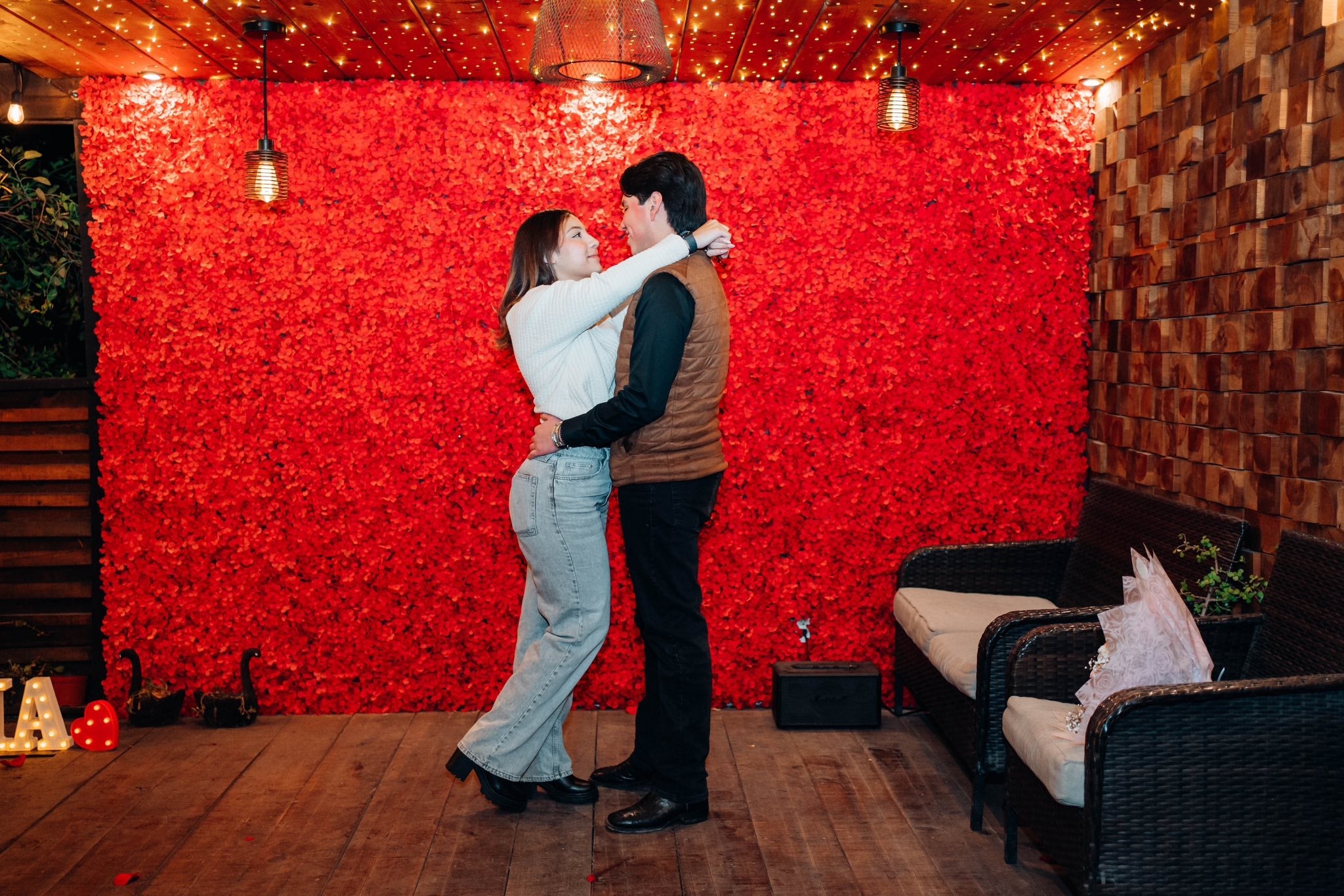 Pareja bailando abrazada frente a un muro de rosas rojas, bajo un techo con luces como estrellas.
