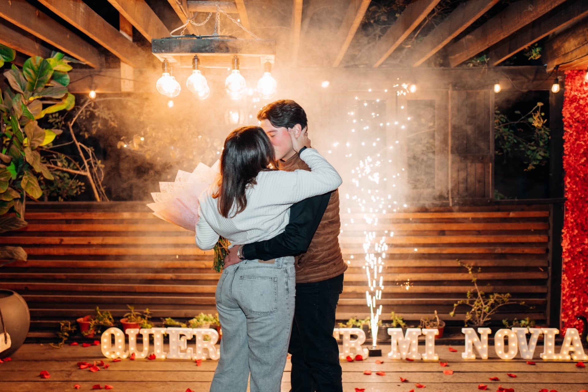Pareja besándose frente a letras luminosas de «Quieres ser mi novia», con pirotecnia fría y humo detrás.